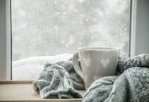 A cozy scene featuring a mug with a heart design and a knitted blanket beside a window, with snow falling softly outside.