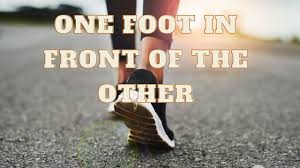 A close-up of a person's foot stepping on a path, with the text 'One Foot In Front Of The Other' overlayed.