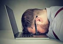 A man in glasses rests his forehead on a laptop, expressing frustration or exhaustion at his work.