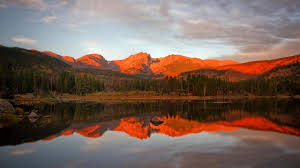 A scenic view of mountains reflecting in a calm lake during sunset, surrounded by trees and a colorful sky.