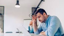 A man in a light blue shirt appears stressed as he sits at a desk, holding his glasses and looking at a laptop. Another person is partially visible in the background.